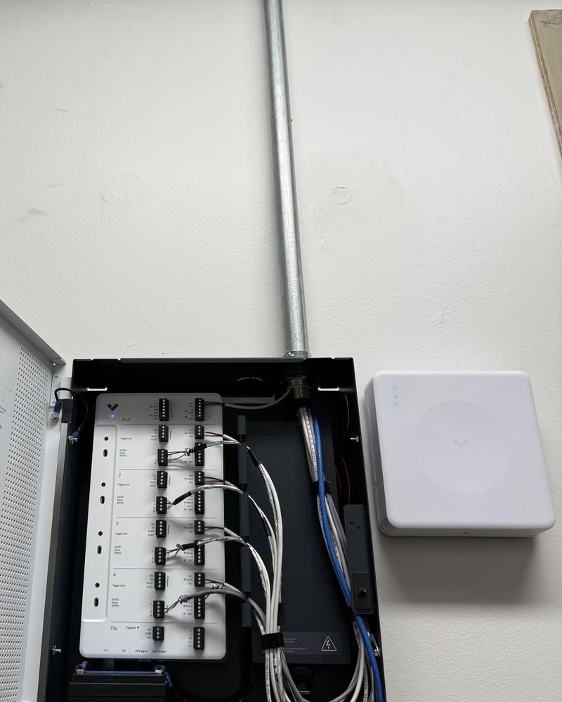 A wall-mounted access control enclosure with labeled terminals and conduit run to the ceiling, beside a ceiling-height wireless access point